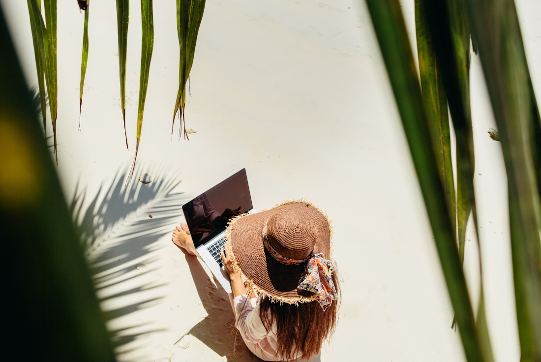 A person sits on a beach with a laptop in their lap.
